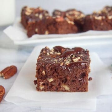 A low carb chocolate pecan brownie topped with nuts is placed on a piece of parchment paper. More brownies can be seen in the background on a white plate.