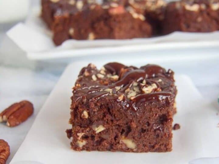 A low carb chocolate pecan brownie topped with nuts is placed on a piece of parchment paper. More brownies can be seen in the background on a white plate.