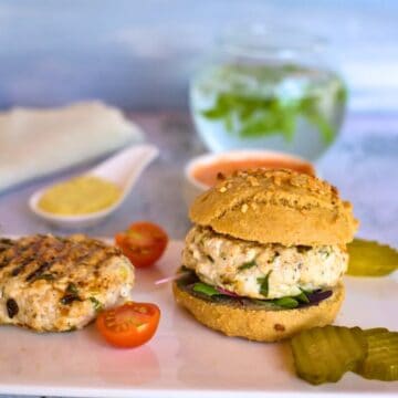 A chicken lemon tarragon burger on a plate with pickles and sliced cherry tomatoes, accompanied by a glass of water with mint leaves in the background.