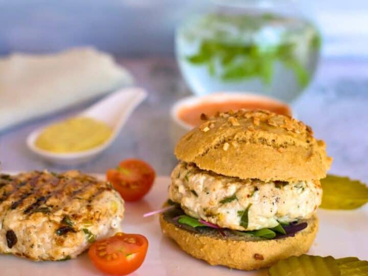 A chicken lemon tarragon burger on a plate with pickles and sliced cherry tomatoes, accompanied by a glass of water with mint leaves in the background.