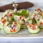 Stuffed cucumber bites filled with a mix of feta cheese, tomatoes, olives, and basil on a white plate.