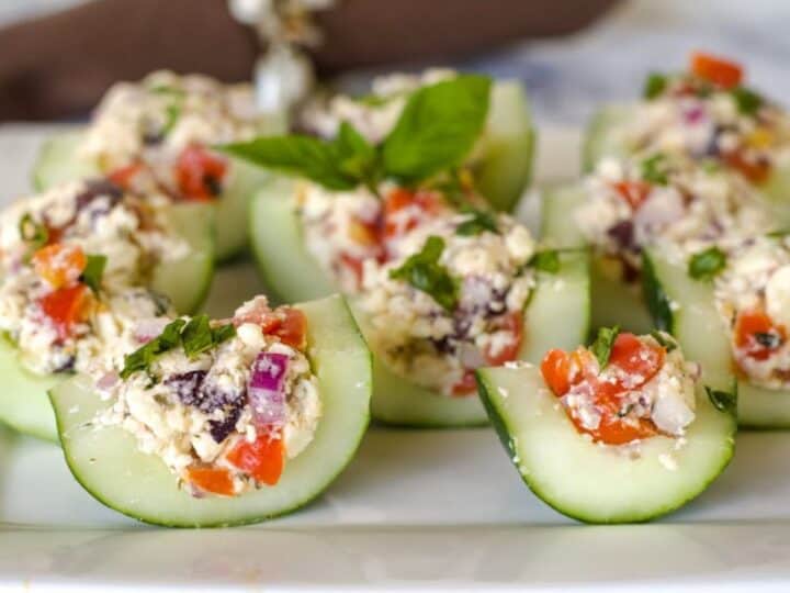 Stuffed cucumber bites filled with a mix of feta cheese, tomatoes, olives, and basil on a white plate.
