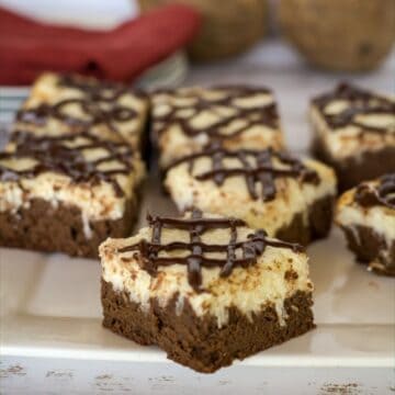 A plate of six keto cheesecake brownies, layered with chocolate and cream and topped with a rich chocolate drizzle, is displayed on a white surface. A red napkin and other objects are blurred in the background.