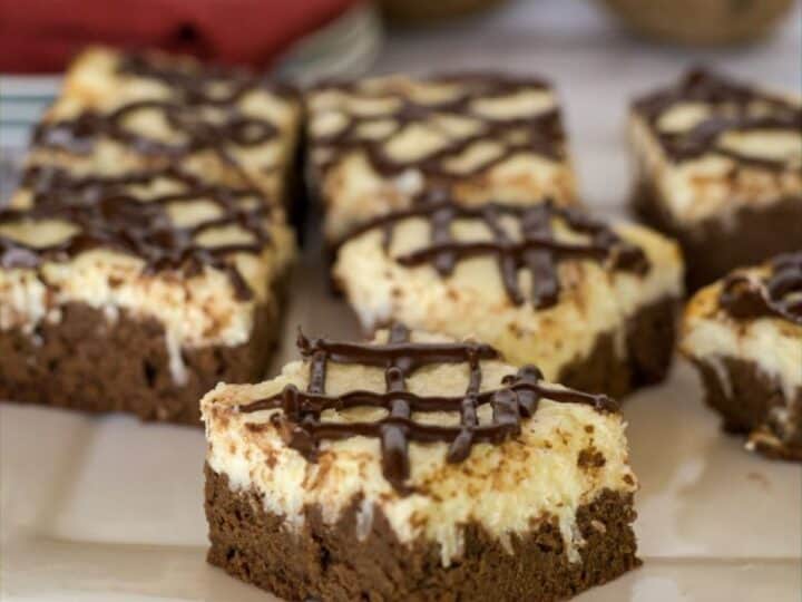 A plate of six keto cheesecake brownies, layered with chocolate and cream and topped with a rich chocolate drizzle, is displayed on a white surface. A red napkin and other objects are blurred in the background.