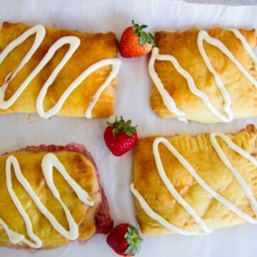 Four rectangular keto danish pastries with a glossy golden-brown finish, drizzled with white icing, are arranged on a white surface. Three fresh strawberries are placed between them.