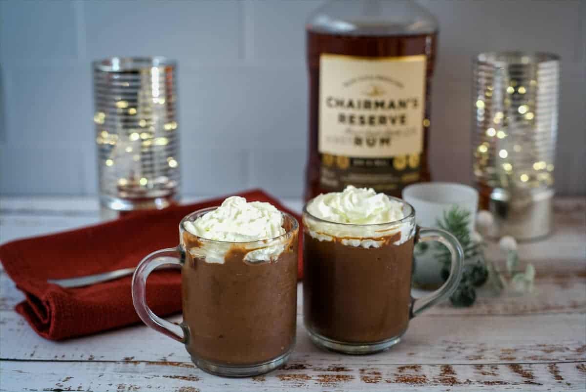 Two glass mugs filled with chocolate rum mousse topped with whipped cream are on a white wooden table. A red napkin, a bottle of rum, and candles in glass holders are in the background.