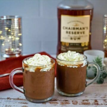 Two glass mugs of hot chocolate topped with whipped cream are on a table. A bottle of rum, candles, a napkin, and holiday decorations are in the background, giving the scene a cozy feel reminiscent of a festive chocolate rum mousse.