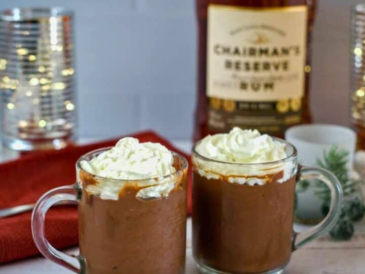 Two glass mugs of hot chocolate topped with whipped cream are on a table. A bottle of rum, candles, a napkin, and holiday decorations are in the background, giving the scene a cozy feel reminiscent of a festive chocolate rum mousse.