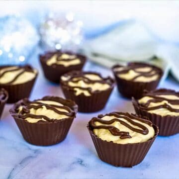 Chocolate cups filled with a low carb peanut butter mousse and topped with chocolate drizzle, arranged on a marble surface with a decorative background.