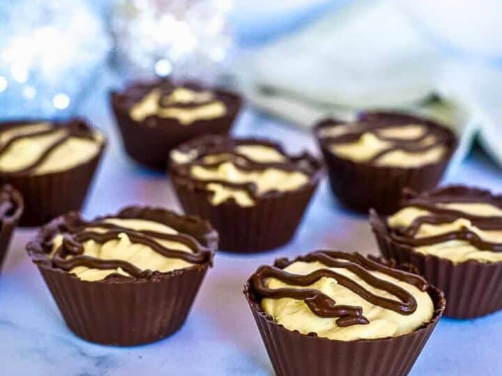 Chocolate cups filled with a low carb peanut butter mousse and topped with chocolate drizzle, arranged on a marble surface with a decorative background.