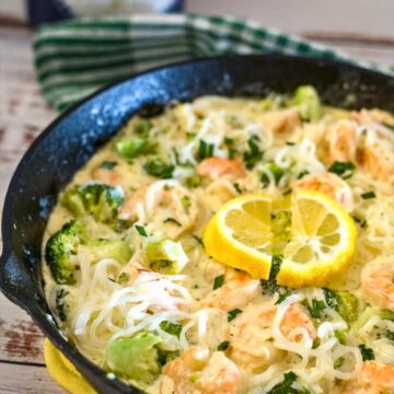 A skillet filled with succulent shrimp broccoli, and noodles in a creamy sauce, garnished with two lemon slices, rests on a wooden surface with a green-striped towel in the background.