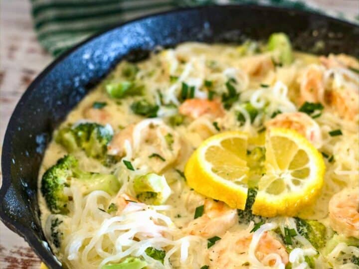 A skillet filled with succulent shrimp broccoli, and noodles in a creamy sauce, garnished with two lemon slices, rests on a wooden surface with a green-striped towel in the background.