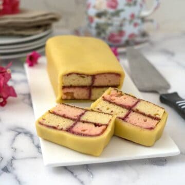 A rectangular Battenberg cake with yellow marzipan, cut to reveal the iconic checkered pink and yellow sponge pattern inside, is displayed on a white plate with a knife next to it.