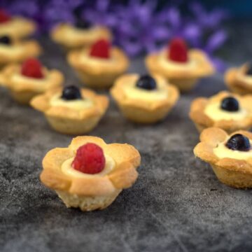 Mini tartlets with flower-shaped crusts, filled with cream and topped with raspberries and blueberries, displayed on a dark surface alongside keto lemon cookies and purple flowers in the background.