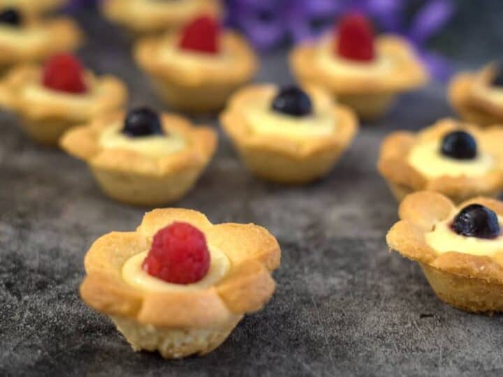 Mini tartlets with flower-shaped crusts, filled with cream and topped with raspberries and blueberries, displayed on a dark surface alongside keto lemon cookies and purple flowers in the background.