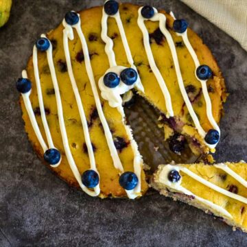 A round Keto Lemon Blueberry Cake with white icing lines and whole blueberries on top, missing one slice. The cake is displayed on a dark surface.