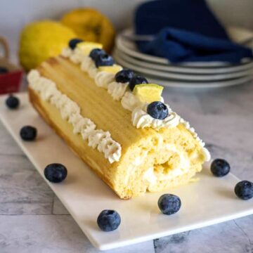 A lemon swiss roll topped with whipped cream, blueberries, and lemon wedges graces a rectangular white plate. Blueberries and a lemon are artfully scattered around, while plates and a napkin add to the elegant setting in the background.