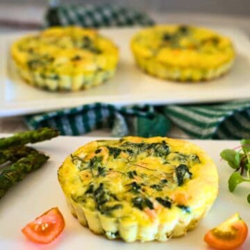 Three spinach and cheese mini quiches are plated alongside asparagus, a halved cherry tomato, and greens. The quiches boast a golden-brown crust while a green-checkered cloth is visible in the background, reminiscent of our delectable crustless salmon quiche.