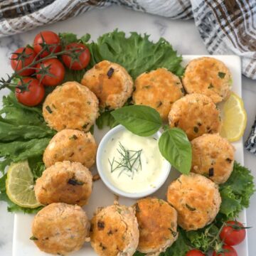 A platter of round keto salmon patties garnished with fresh greens, cherry tomatoes, and lemon slices, centered around a small bowl of creamy dipping sauce with a basil leaf garnish.