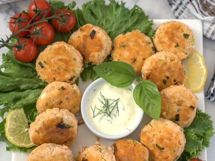 A platter of round keto salmon patties garnished with fresh greens, cherry tomatoes, and lemon slices, centered around a small bowl of creamy dipping sauce with a basil leaf garnish.