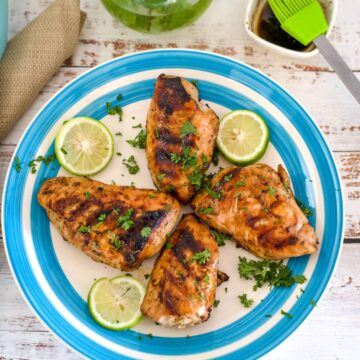 A white and blue plate with four grilled keto BBQ chicken breasts garnished with chopped herbs and lime slices. A sauce brush and a bowl of sauce are partially visible in the background.