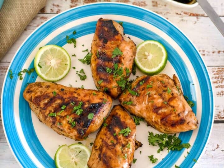 A white and blue plate with four grilled keto BBQ chicken breasts garnished with chopped herbs and lime slices. A sauce brush and a bowl of sauce are partially visible in the background.