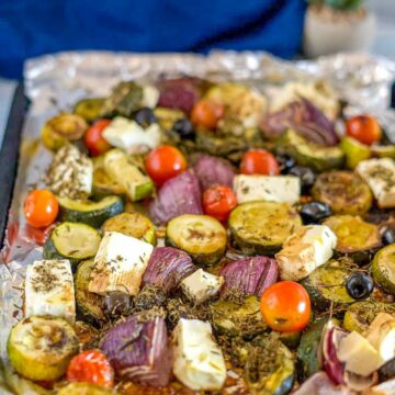 A baking sheet with roasted, seasoned vegetables, including zucchini, red onion, cherry tomatoes, olives, and blocks of feta cheese—a perfect prelude to a zucchini tomato salad.