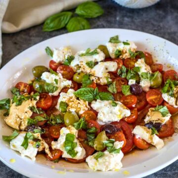 A plate of fresh Caprese salad with cherry tomatoes, mozzarella, green and black olives, basil leaves, and a drizzle of olive oil brings together the essence of a tomato burrata salad.