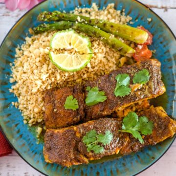 A plate with two pieces of Moroccan salmon garnished with cilantro, served with couscous and asparagus, and topped with lime slices.