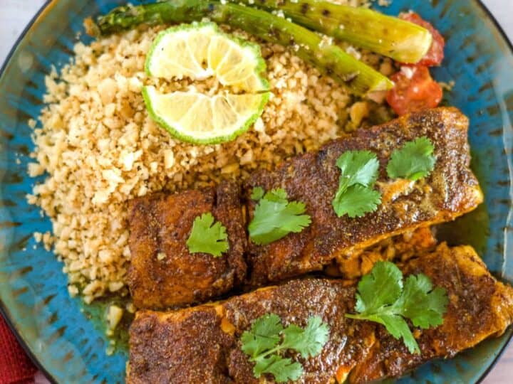 A plate with two pieces of Moroccan salmon garnished with cilantro, served with couscous and asparagus, and topped with lime slices.