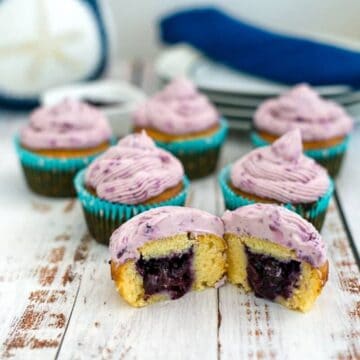 Keto blueberry cupcakes with purple frosting and blue liners are arranged on a wooden surface. One cupcake is cut open, displaying a delightful blueberry filling inside. Plates and a napkin are visible in the background.