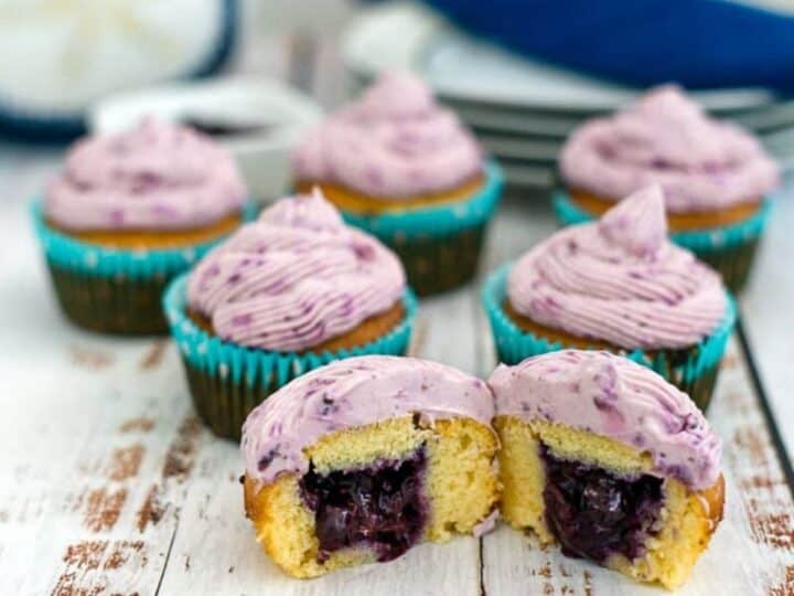 Keto blueberry cupcakes with purple frosting and blue liners are arranged on a wooden surface. One cupcake is cut open, displaying a delightful blueberry filling inside. Plates and a napkin are visible in the background.