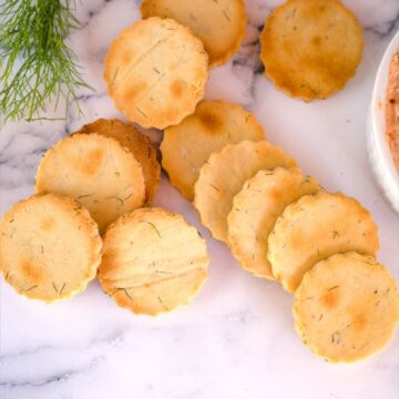 A group of round, golden-brown, scalloped-edged almond flour crackers are arranged on a marble surface next to a small bunch of greens.