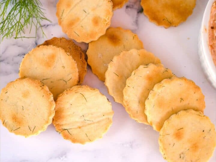 A group of round, golden-brown, scalloped-edged almond flour crackers are arranged on a marble surface next to a small bunch of greens.