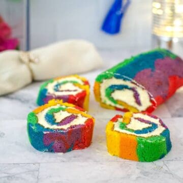 Three slices of a colorful rainbow cake roll with swirls of cream filling are arranged on a marble surface alongside the remaining unsliced roll.