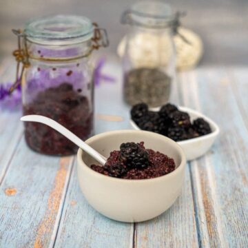 A bowl of chia seed pudding topped with blackberries sits next to a similar bowl brimming with them, while a jar filled with blackberry chia jam stands invitingly in the background.