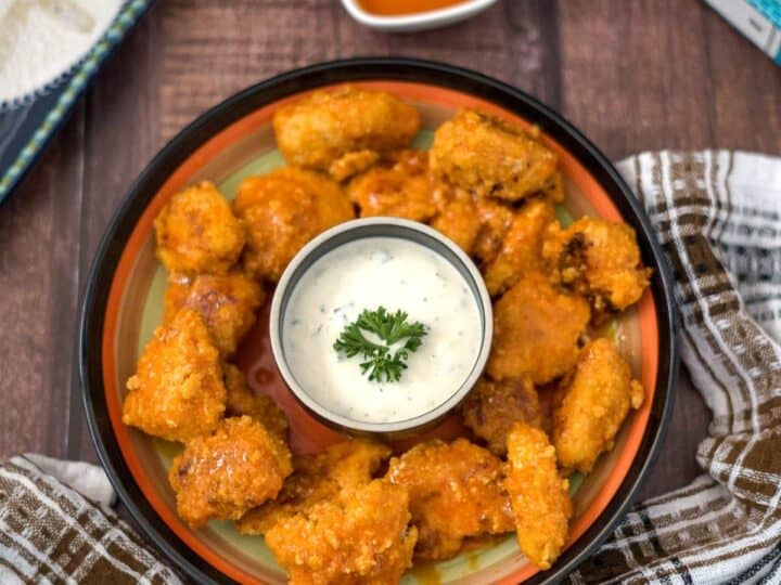 A plate of buffalo chicken bites served with a small bowl of dipping sauce, garnished with a sprig of parsley. There is a napkin on the side and a separate sauce bowl in the background.