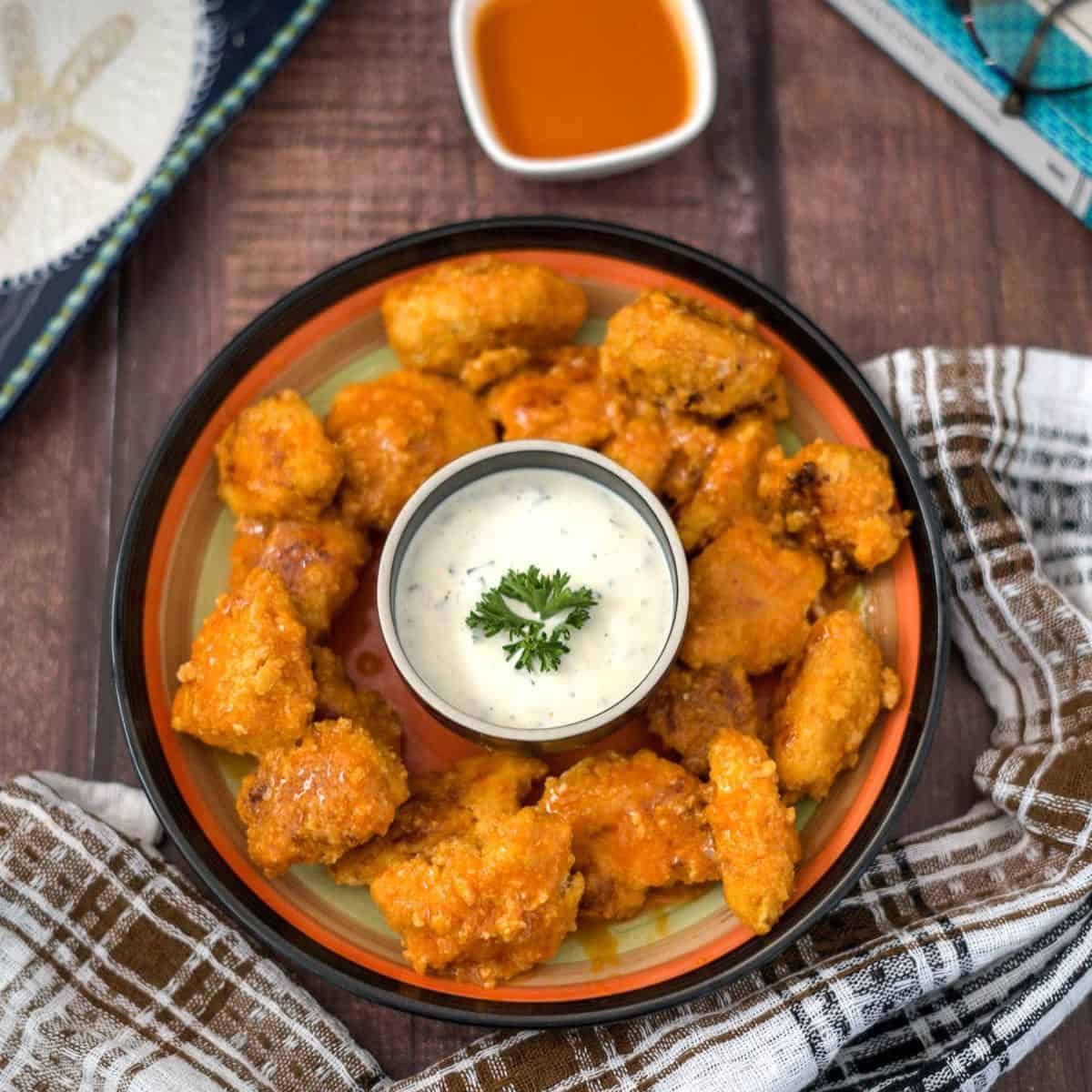 A plate of buffalo chicken bites served with a small bowl of dipping sauce, garnished with a sprig of parsley. There is a napkin on the side and a separate sauce bowl in the background.