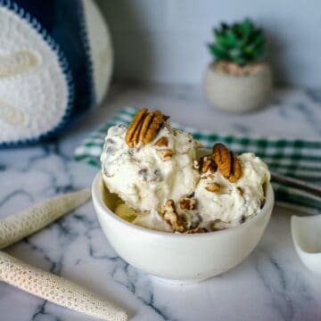 A bowl of keto butter pecan ice cream is placed on a marble table, topped with whole pecans. The background features a green and white cloth, a plant, a decorative object, and two starfish ornaments.