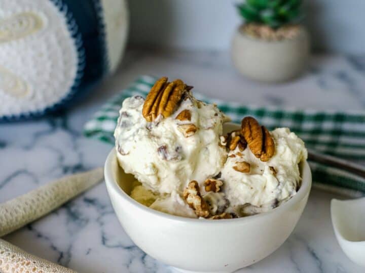 A bowl of keto butter pecan ice cream is placed on a marble table, topped with whole pecans. The background features a green and white cloth, a plant, a decorative object, and two starfish ornaments.