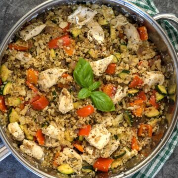 A large pan filled with a cooked mixture of chicken pieces, diced red bell peppers, zucchini, onions, and Mediterranean cauliflower rice, garnished with a sprig of basil on top.