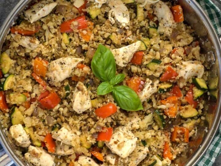 A large pan filled with a cooked mixture of chicken pieces, diced red bell peppers, zucchini, onions, and Mediterranean cauliflower rice, garnished with a sprig of basil on top.