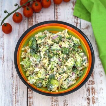 A colorful bowl filled with a broccoli ranch salad, garnished with sunflower seeds and accompanied by a bunch of cherry tomatoes and a green napkin on a wooden surface.