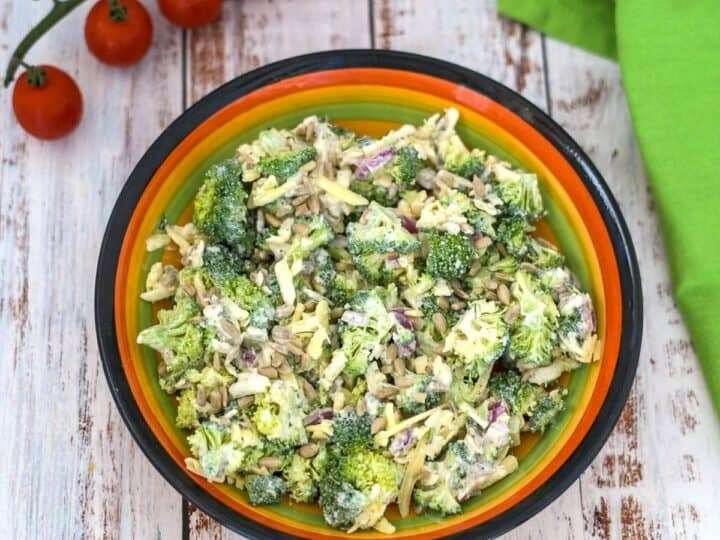 A colorful bowl filled with a broccoli ranch salad, garnished with sunflower seeds and accompanied by a bunch of cherry tomatoes and a green napkin on a wooden surface.