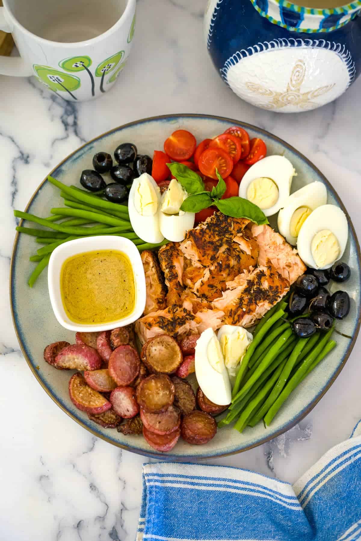 A plate showcasing a keto salmon salad with green beans, boiled eggs, cherry tomatoes, black olives, roasted potatoes, and mustard sauce. A mug and a jug are in the background, with a cloth napkin beside the plate.