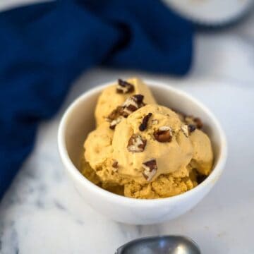 A white bowl filled with scoops of pumpkin cheesecake ice cream topped with chopped pecans, placed on a marble surface with a blue napkin in the background.