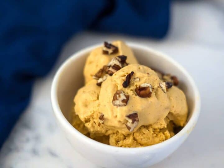 A white bowl filled with scoops of pumpkin cheesecake ice cream topped with chopped pecans, placed on a marble surface with a blue napkin in the background.