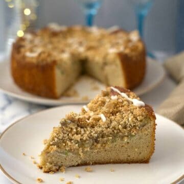 A slice of crumb-topped keto apple cake served on a white plate, with the rest of the cake on a platter in the background.