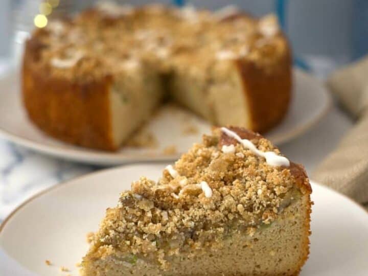 A slice of crumb-topped keto apple cake served on a white plate, with the rest of the cake on a platter in the background.