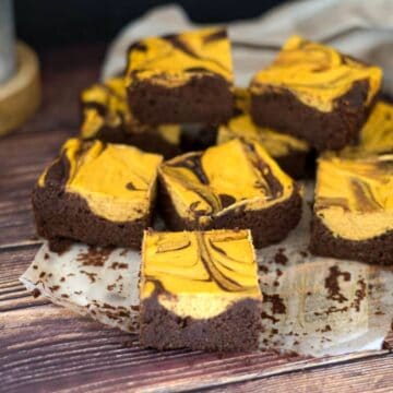 A batch of chocolate and pumpkin cheesecake swirl brownies arranged on a parchment paper atop a wooden surface.